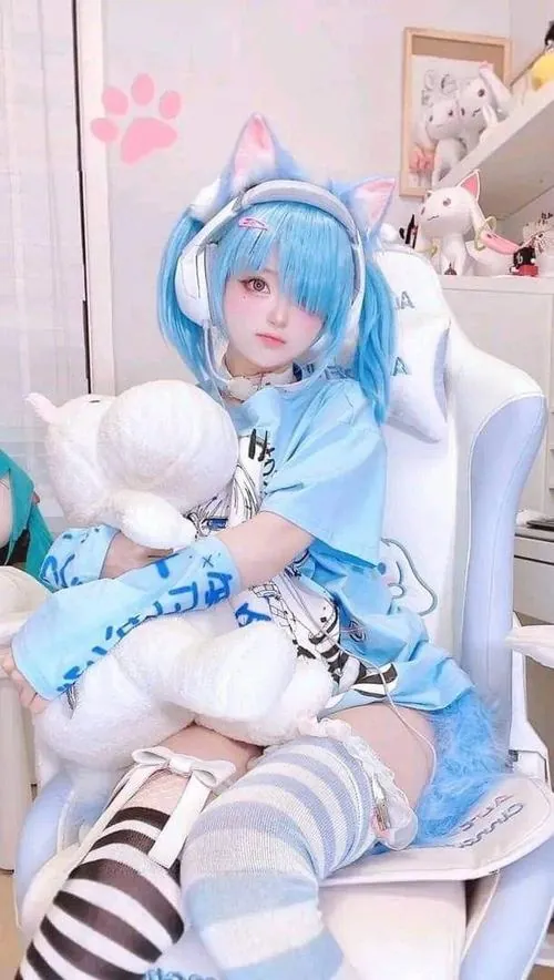 Cô gái tóc xanh ôm thú bông trong không gian trong nhà, trang phục cosplay sáng màu làm khung hình thêm dịu mắt.
