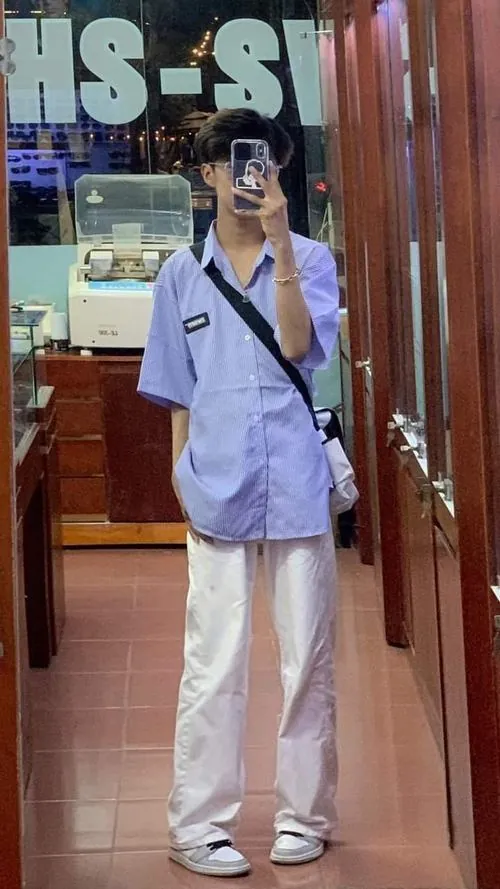 Bạn nam mặc áo sơ mi xanh nhạt và quần trắng đứng trong nhà, dáng chụp cắt mặt tạo vẻ tự nhiên.
