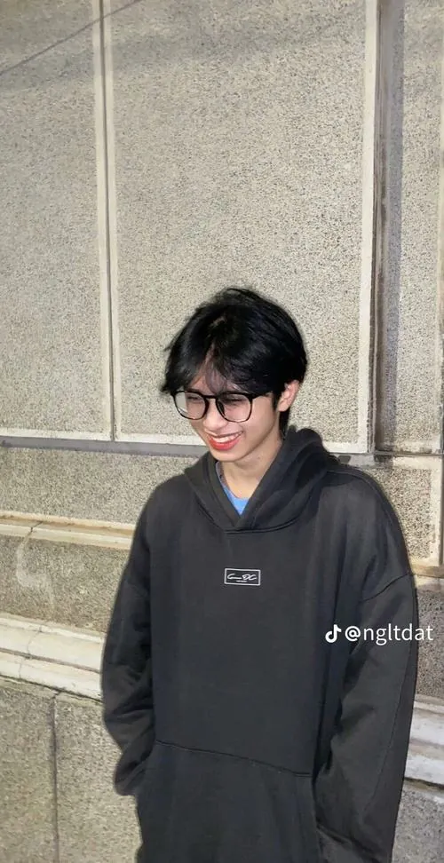 Ảnh trai k12 mặc hoodie đen, đeo kính và cúi cười bên bức tường đá, tạo cảm giác gần gũi của một buổi tan học.