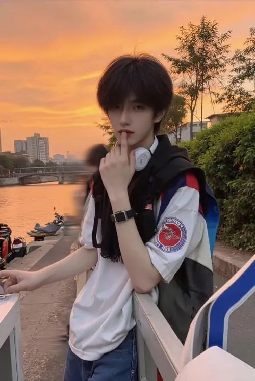 Nam sinh ngồi bên lan can lúc hoàng hôn, áo trắng nổi bật giữa nền trời cam, ánh nhìn nghiêng gợi chút lặng lẽ cuối ngày.