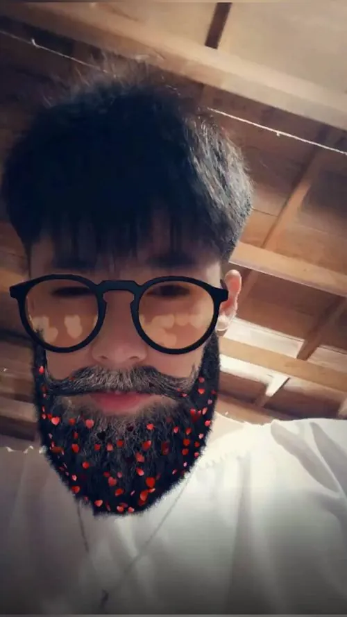 Bạn nam đeo kính tròn dùng filter râu, ngồi trong nhà với ánh đèn ấm tạo một khoảnh khắc selfie hài hước.