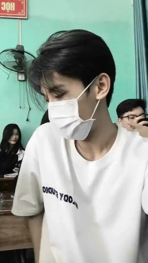 Học sinh nam mặc áo phông trắng, đeo khẩu trang và nhìn sang bên trong lớp, phía sau có vài bạn đang ngồi học.