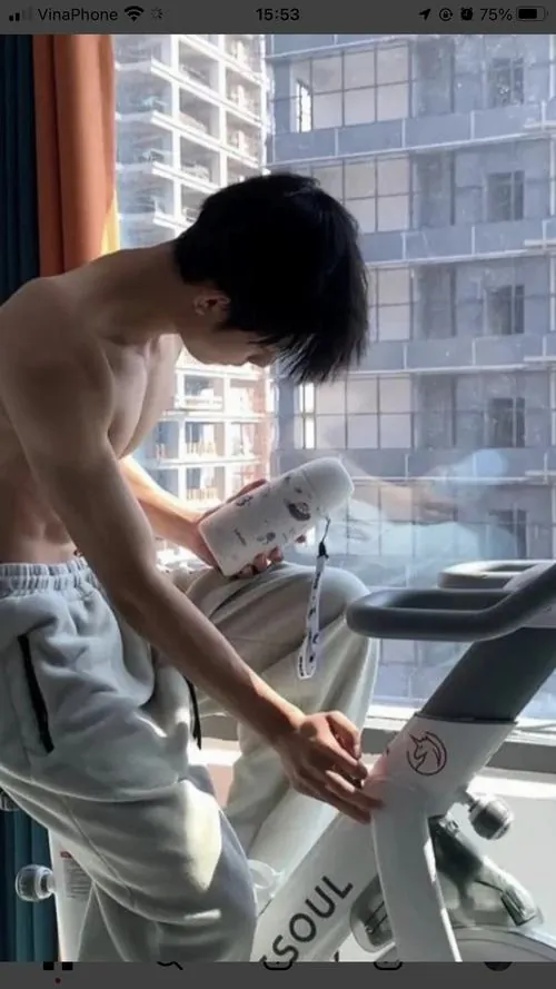 Chàng trai cúi người bên máy chạy, lưng và vai hiện rõ dưới ánh cửa sổ, bối cảnh phòng tập sáng và khá yên tĩnh.