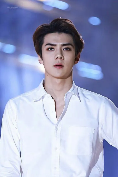 Người mẫu nam mặc sơ mi trắng ngồi trên bục xi măng trong sân thể thao vắng, cổ áo hơi mở sau buổi tập.