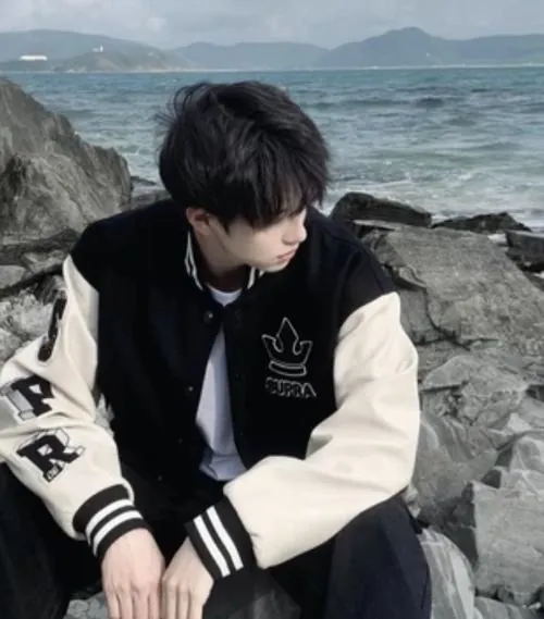 Nam sinh ngồi trên ghềnh đá ven biển, áo khoác varsity đen trắng và dáng cúi đầu khiến ảnh mang cảm giác lặng lại.