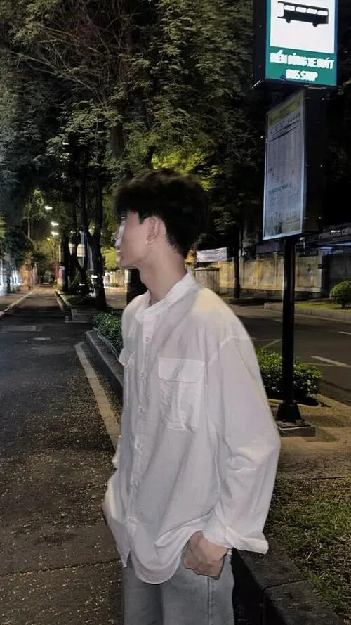 Chàng trai tóc đen khoác sơ mi trắng đứng nghiêng góc phố ban đêm, tay vuốt nhẹ mái tóc che đi nửa mặt dưới ánh đèn đường vàng nhạt.
