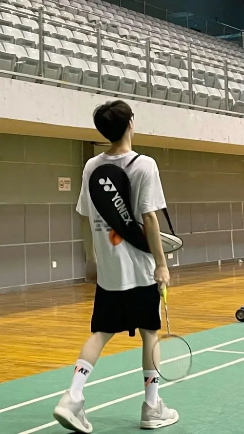 Chàng trai đeo bao vợt cầu lông sau lưng, khoác áo phông trắng năng động bước đi trong nhà thi đấu với vóc dáng cao ráo.
