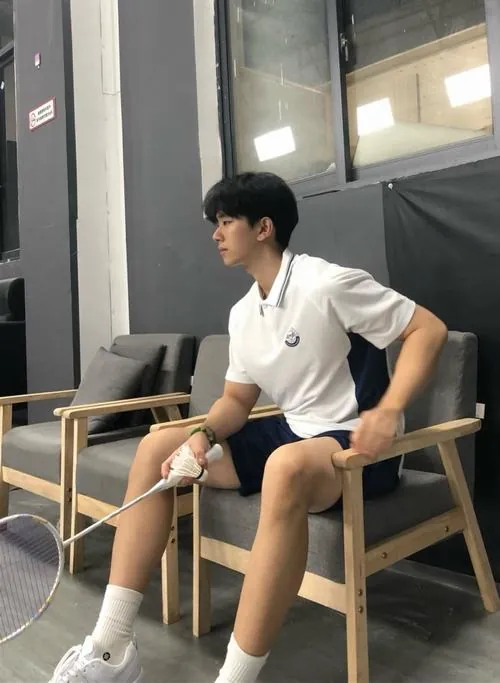 Hot boy cầu lông ngồi thư giãn trên chiếc ghế bành trắng, tay cầm chai nước khoáng giải khát sau trận đấu căng thẳng.
