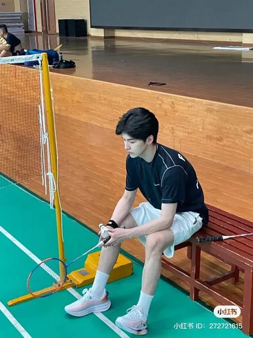 Hot boy sân cầu ngồi nghỉ trên băng ghế gỗ, tay cầm vợt miết nhẹ mặt lưới với ánh mắt chăm chú đầy tập trung.