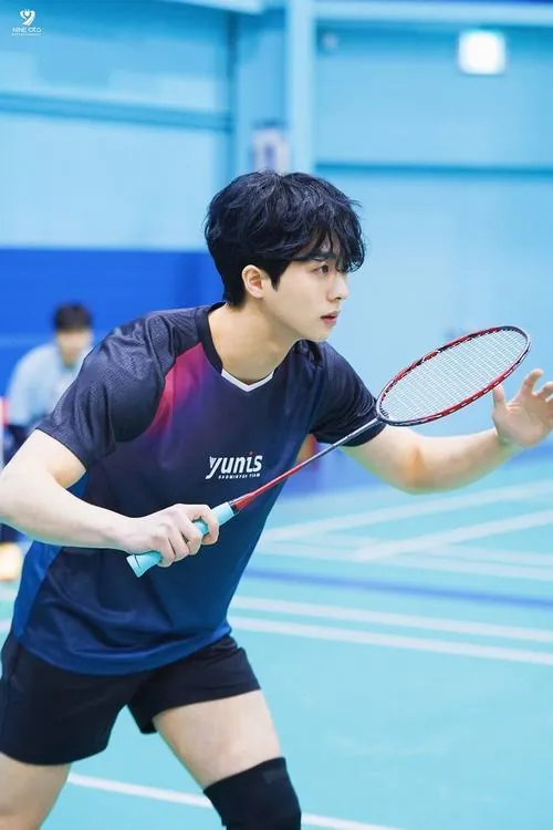 Tay vợt trẻ đứng tựa vào cột lưới ngoài trời, vừa mỉm cười rạng rỡ vừa đưa tay che ánh nắng mặt trời chiếu qua kẽ lá.